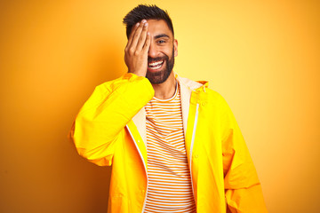 Young indian man wearing raincoat standing over isolated yellow background covering one eye with hand, confident smile on face and surprise emotion.