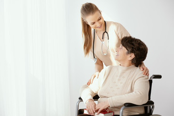 Young smiling doctor in beige uniform with senior patient on wheelchair, photo with copy space on...