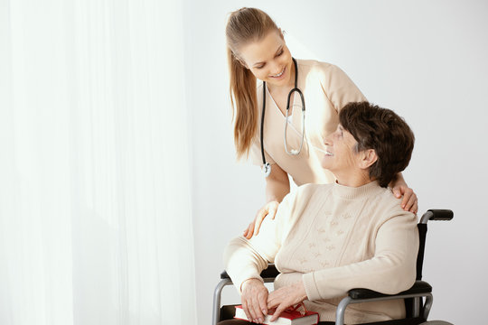 Young smiling doctor in beige uniform with senior patient on wheelchair, photo with copy space on the empty wall
