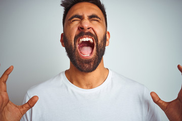 Young indian man wearing t-shirt standing over isolated white background crazy and mad shouting and yelling with aggressive expression and arms raised. Frustration concept.