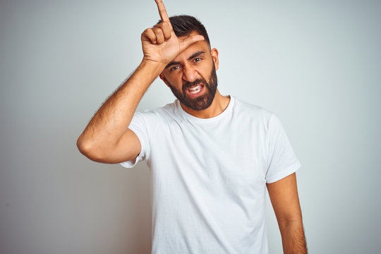 Young indian man wearing t-shirt standing over isolated white background making fun of people with fingers on forehead doing loser gesture mocking and insulting.