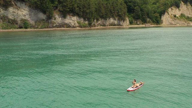 Sup surfer woman rowing on supboard. Paddle board sailing on turquoise water along beautiful green cliff. Beautiful seascape. Man resting lying on surfboard. Outdoor summer water sport aerial view
