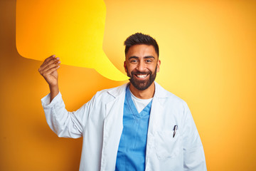 Young indian doctor man holding speech bubble standing over isolated yellow background with a happy face standing and smiling with a confident smile showing teeth