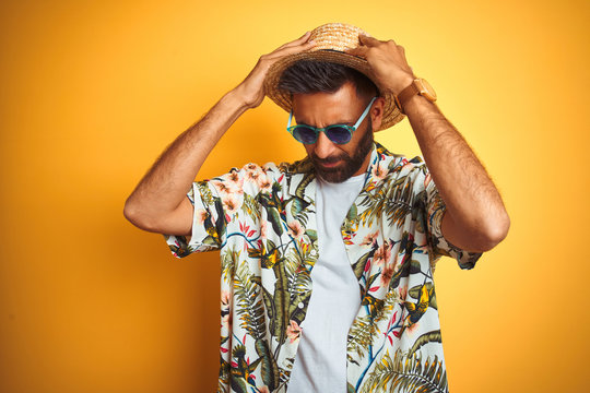 Indian man on vacation wearing floral shirt hat sunglasses over isolated yellow background suffering from headache desperate and stressed because pain and migraine. Hands on head.