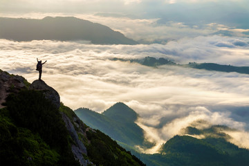 Wide mountain panorama. Small silhouette of tourist with backpack on rocky mountain slope with raised hands over valley covered with white puffy clouds. Beauty of nature, tourism and traveling concept