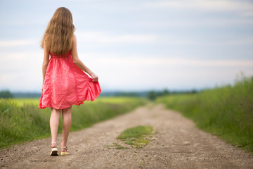 Back view of young romantic slim woman in red dress with long hair walking by ground road along green field on sunny summer day on blue sky copy space background.