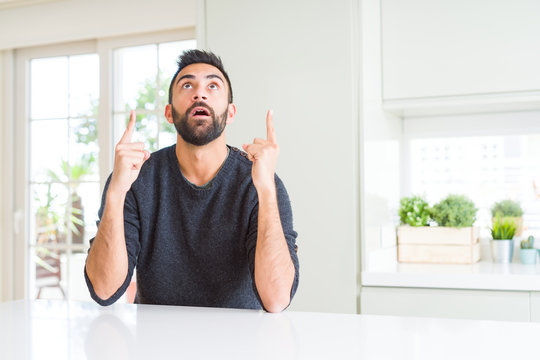 Handsome hispanic man wearing casual sweater at home amazed and surprised looking up and pointing with fingers and raised arms.