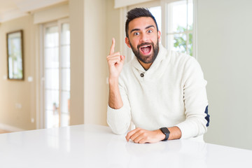 Handsome hispanic man wearing casual white sweater at home pointing finger up with successful idea. Exited and happy. Number one.