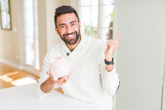 Handsome hispanic man holding piggy bank very happy pointing with hand and finger to the side