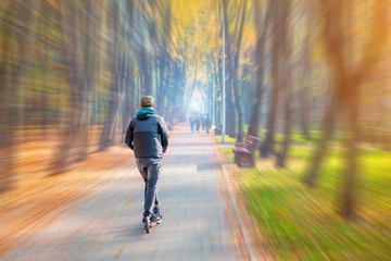 Young adult person riding modern electric scooter along beautiful colorful autumn city park. Man driving gadget vehicle through multicolored fall tree valley. Motion blur
