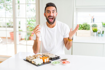 Handsome hispanic man eating asian sushi using chopsticks very happy and excited, winner expression celebrating victory screaming with big smile and raised hands