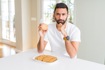 Handsome hispanic man eating healthy whole grain biscuits serious face thinking about question,...