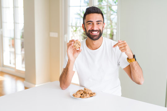 Handsome hispanic man eating chocolate chips cookies with surprise face pointing finger to himself