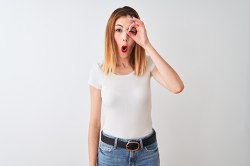 Beautiful redhead woman wearing casual t-shirt standing over isolated white background doing ok gesture shocked with surprised face, eye looking through fingers. Unbelieving expression.