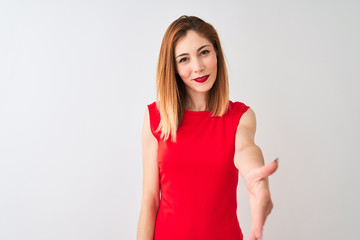Redhead businesswoman wearing elegant red dress standing over isolated white background smiling friendly offering handshake as greeting and welcoming. Successful business.
