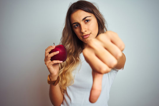 Young beautiful woman eating red apple over grey isolated background with angry face, negative sign showing dislike with thumbs down, rejection concept