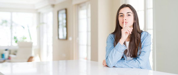 Wide angle picture of beautiful young woman sitting on white table at home asking to be quiet with finger on lips. Silence and secret concept.