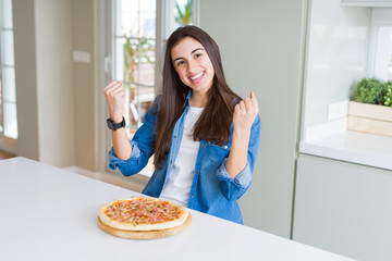 Beautiful young woman eating homemade tasty pizza at the kitchen celebrating surprised and amazed for success with arms raised and open eyes. Winner concept.