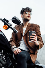 low angle view of stylish man in jacket and sunglasses holding coffee to go while sitting on motorcycle