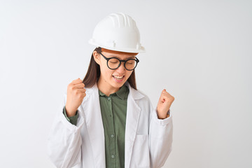 Young chinese engineer woman wearing coat helmet glasses over isolated white background very happy and excited doing winner gesture with arms raised, smiling and screaming for success. 