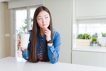 Beautiful Asian woman drinking a fresh glass of milk serious face thinking about question, very confused idea