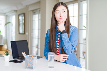 Beautiful Asian woman working using computer laptop with hand on chin thinking about question, pensive expression. Smiling with thoughtful face. Doubt concept.