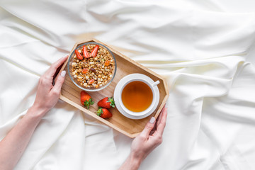 Breakfast on the tray in hands with granola, tea and fruit on white bed sheet background top view