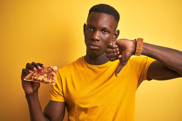 African american man eating slice of pizza standing over isolated yellow background with angry...