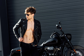 Fototapeta na wymiar young man with muscular torso in leather jacket standing near black motorcycle