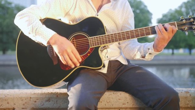 Man sits on a stone boardwalk in the park and plays the guitar. Close up shot.