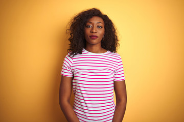 Young african american woman wearing striped t-shirt over isolated yellow background with serious expression on face. Simple and natural looking at the camera.