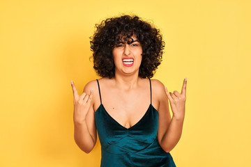 Young arab woman with curly hair wearing elegant dress over isolated yellow background shouting with crazy expression doing rock symbol with hands up. Music star. Heavy concept.