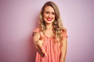 Young beautiful woman wearing t-shirt standing over pink isolated background smiling friendly offering handshake as greeting and welcoming. Successful business.