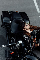Fototapeta na wymiar young man in glasses and brown jacket resting while sitting on ground and leaninghead on motorcyce