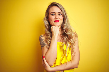 Young attactive woman wearing t-shirt standing over yellow isolated background looking confident at the camera with smile with crossed arms and hand raised on chin. Thinking positive.