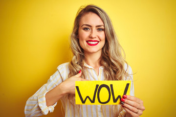 Beautiful woman holding amazed wow surprise banner over isolated yellow background with surprise face pointing finger to himself