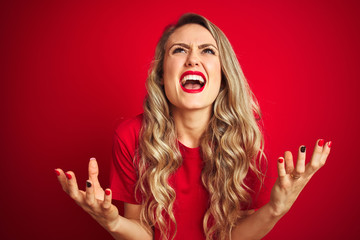 Young beautiful woman wearing basic t-shirt standing over red isolated background crazy and mad shouting and yelling with aggressive expression and arms raised. Frustration concept.