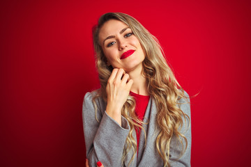 Young beautiful business woman wearing elegant jacket standing over red isolated background looking confident at the camera smiling with crossed arms and hand raised on chin. Thinking positive.