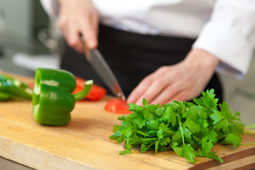 A person professional cook in uniform in the kitchen cuts lettuce (tomatoes) on a wooden board. Closeup of hands, knife, green parsley and pepper. The background is blurred
