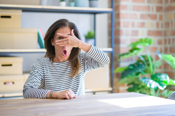 Middle age senior woman sitting at the table at home peeking in shock covering face and eyes with hand, looking through fingers with embarrassed expression.