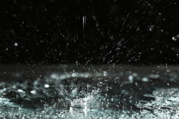 Rain drop falling down into puddle on dark background