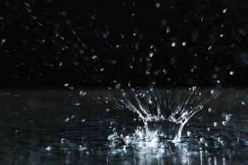 Rain drop falling down into puddle on dark background