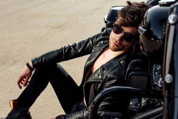 handsome young man in sunglasses leaning on motorcycle while sitting on ground