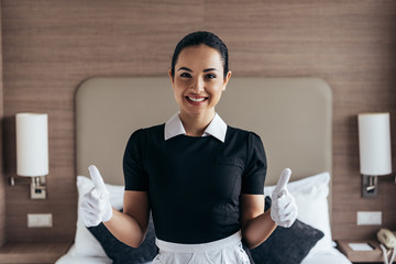 front view of pretty smiling maid in white gloves and apron looking at camera and showing thumbs up in hotel room - obrazy, fototapety, plakaty