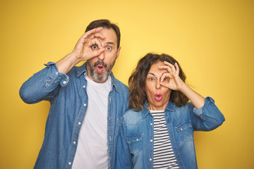Beautiful middle age couple together wearing denim shirt over isolated yellow background doing ok gesture shocked with surprised face, eye looking through fingers. Unbelieving expression.