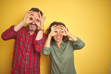 Beautiful middle age couple over isolated yellow background doing ok gesture like binoculars sticking tongue out, eyes looking through fingers. Crazy expression.