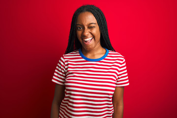 Young african american woman wearing striped t-shirt standing over isolated red background winking...