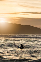 Surfer woman waiting for the perfect wave sitting on her surfboard with sunset light in northern Spain