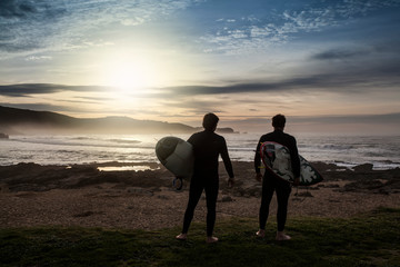 two unrecognizable young man surfers walking with their boards on the rocks to enter the sea to ride waves, seen from behind at sunset