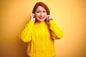 Beautiful redhead woman wearing winter sweater standing over isolated yellow background covering ears with fingers with annoyed expression for the noise of loud music. Deaf concept.